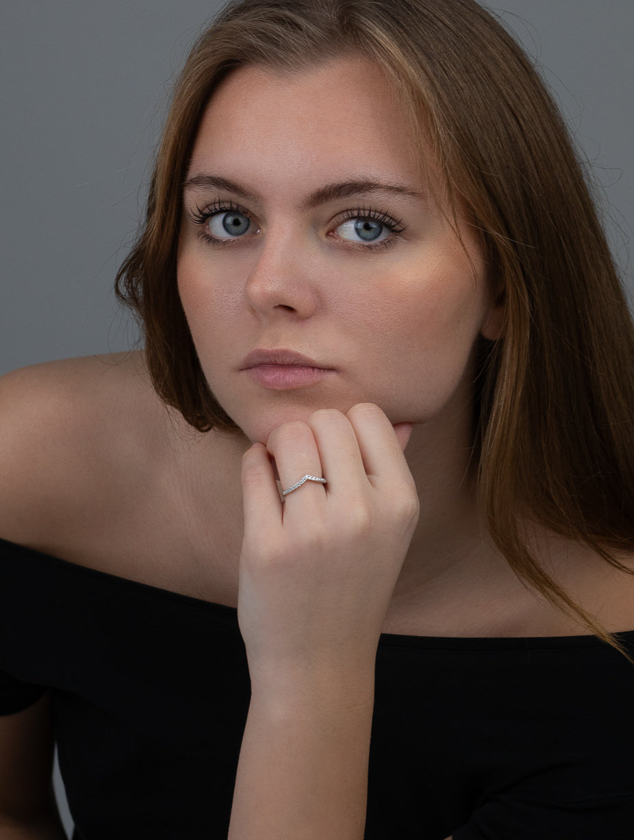 En modell som bär en smal v-formad ring i silver. Modellen tittar rakt fram med handen mot ansiktet.