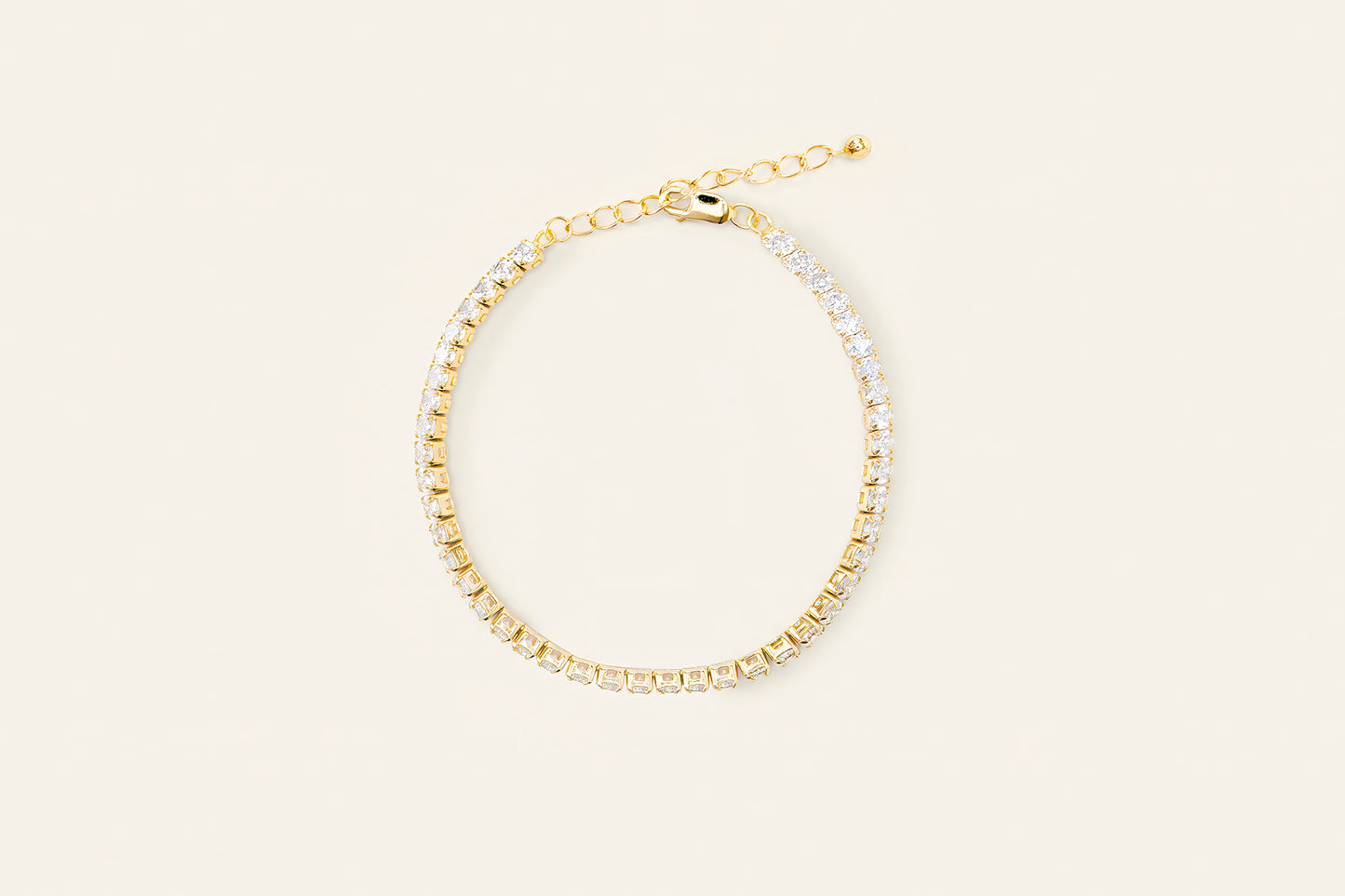 Ett tennisarmband i guld täckt med silver zirconia stenar. Guldarmbandet är placerat på en ljus beige bakgrund.